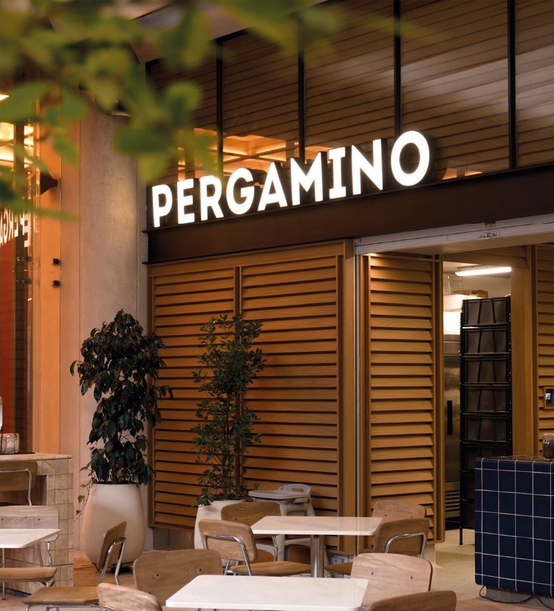 Pergamino: la fachada del café dentro de The Cut, con el letrero iluminado y mesas afuera.