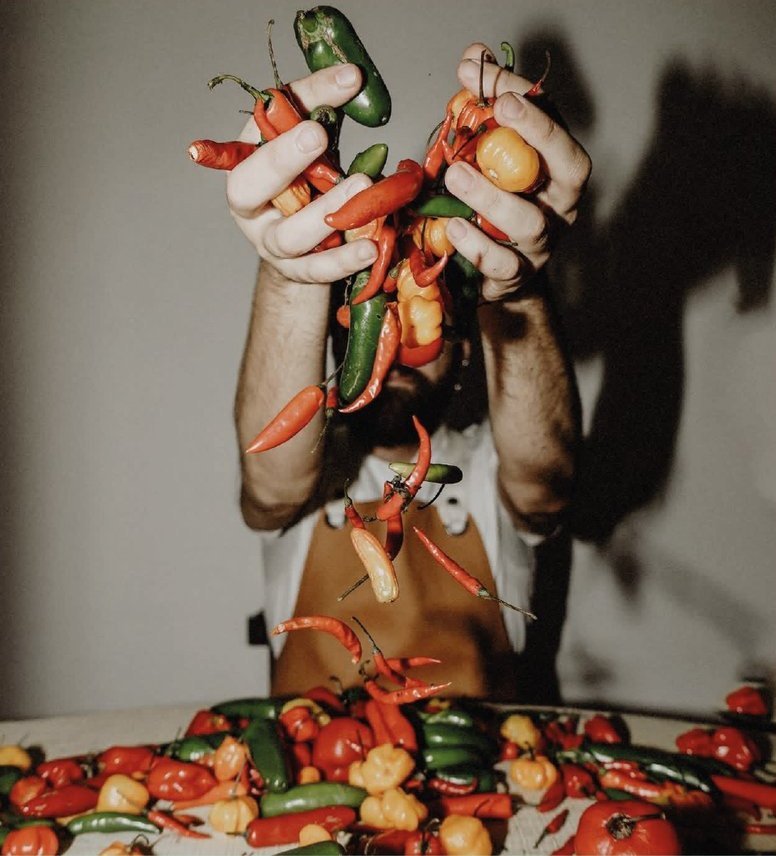 Zombra: dos manos llenas de ajíes rojos y verdes sobre una mesa cubierta de ajíes y tomates.