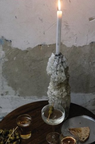 Persona Sala: una vela encendida sobre un candelabro de cera escurrida, en una mesa redonda con copas y aceitunas.