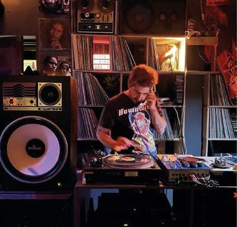 Siete Pulgadas: alguien poniendo un vinilo frente a una pared de discos y un parlante grande.